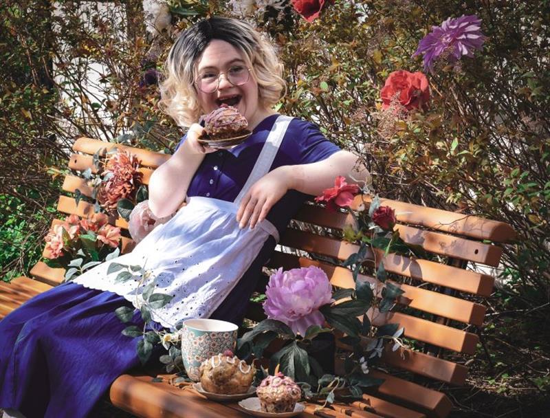 Alice i Eventyrland_web_Foto_Siraffen kultur og kompetansesenter.jpg En jente som sitter på en benk og spiser bakst. Rundt henne er det planter og blomster.