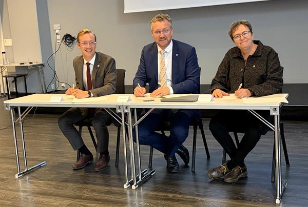 To menn og en kvinne sitter ved et bord og skriver under en erklæring